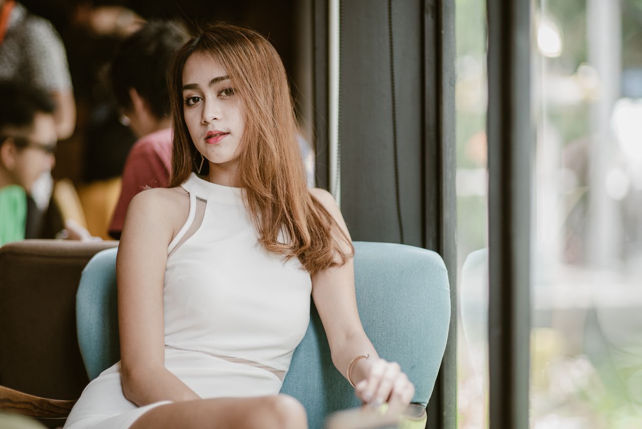 A woman in a white dress sitting by a window indoors with a relaxed and thoughtful pose.