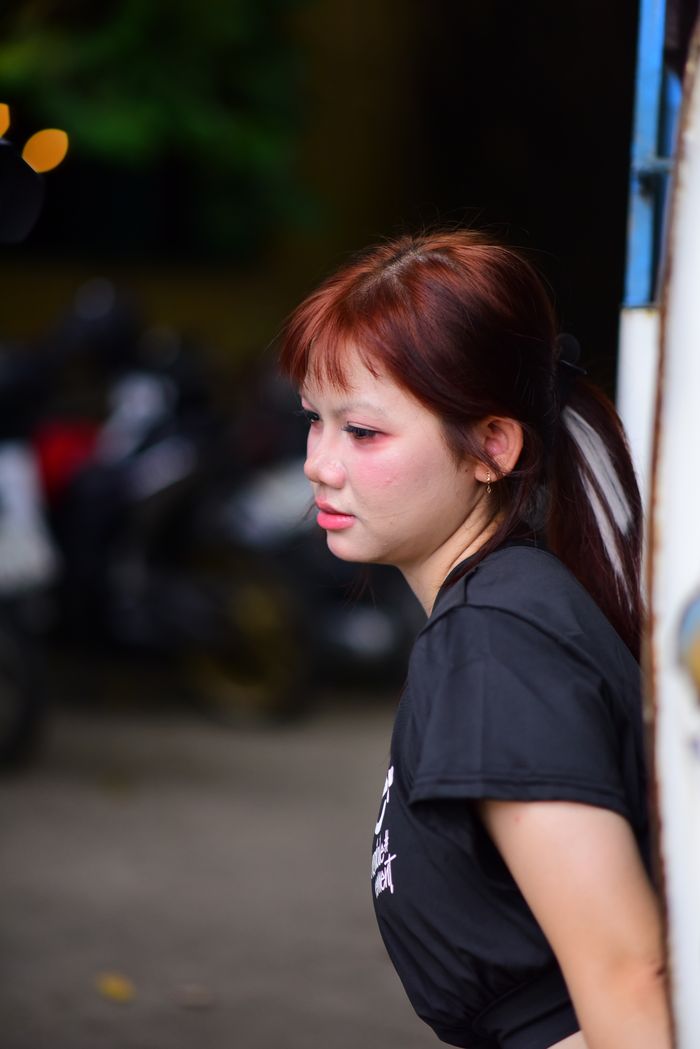 Pensive young woman with red hair stands leaning against a wall outdoors.
