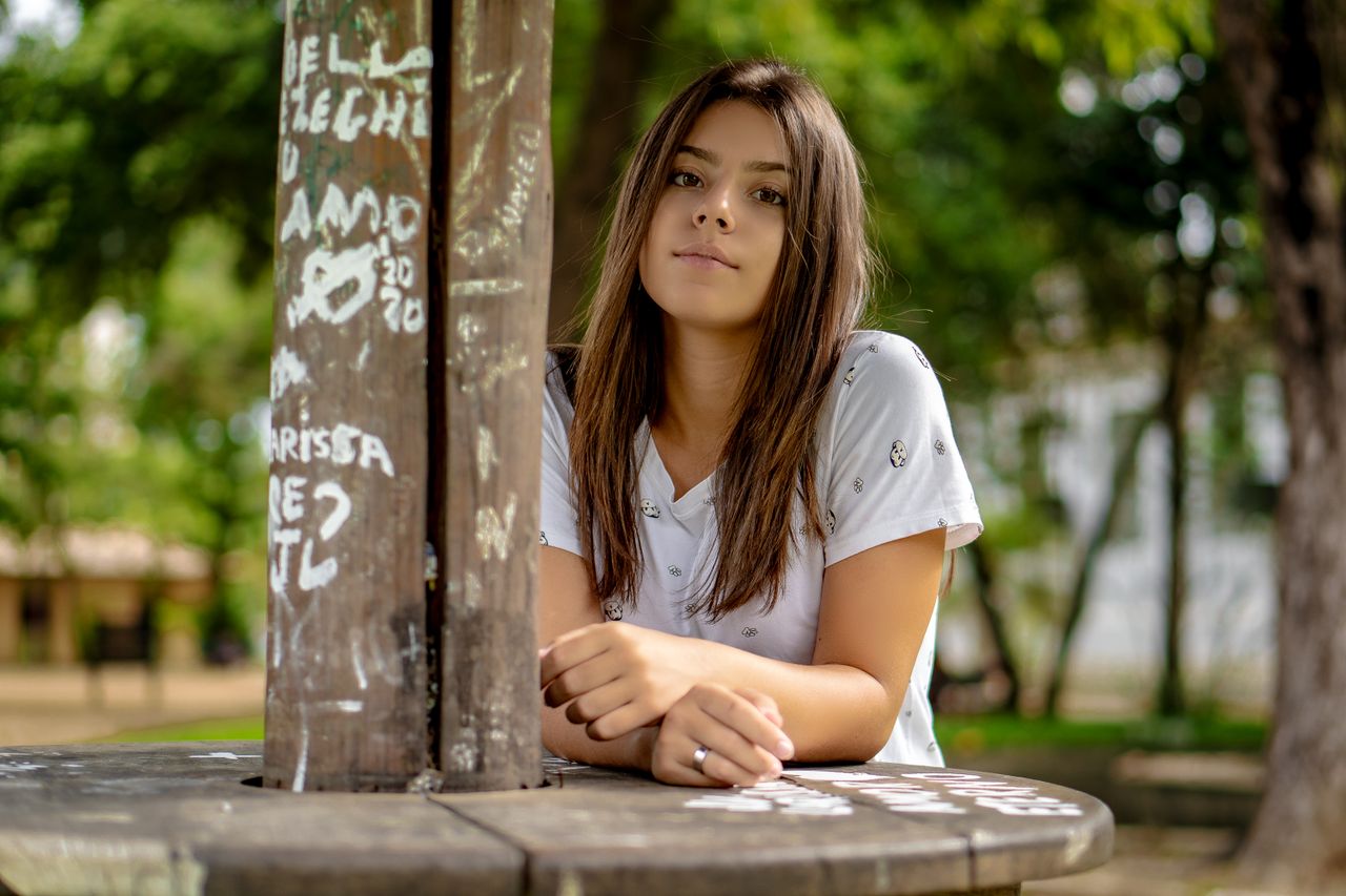 A young woman with long brunette hair poses confidently in an outdoor park setting with graffiti decor.