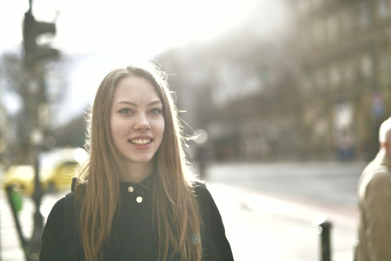 A young woman smiling brightly outdoors on a sunny street.