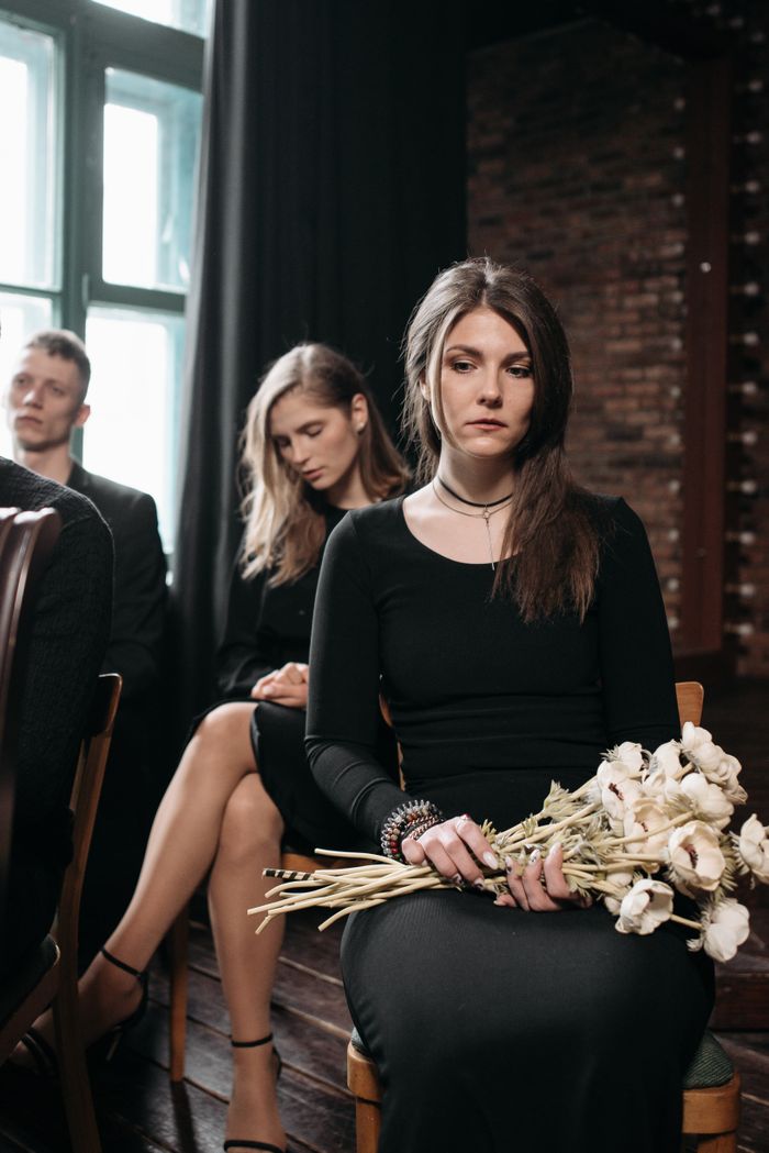 A group of people attend a somber event indoors, with a woman in a black dress holding white roses.